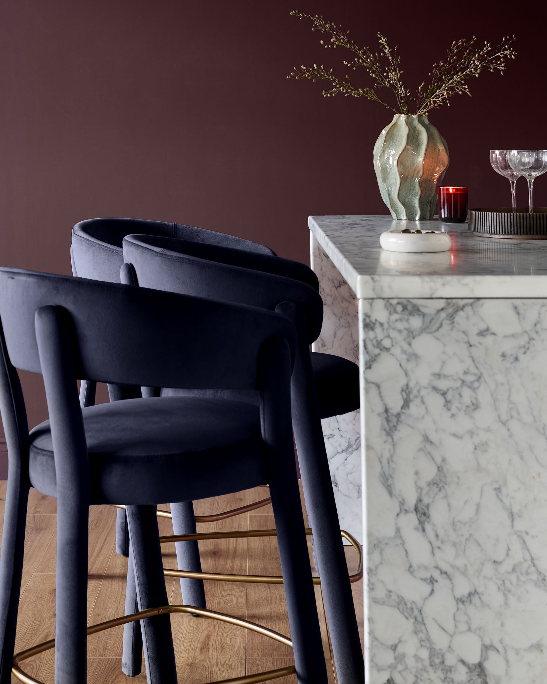 Anya bar stool in dark navy velvet with curved armrests and brass footrest, shown at marble counter.