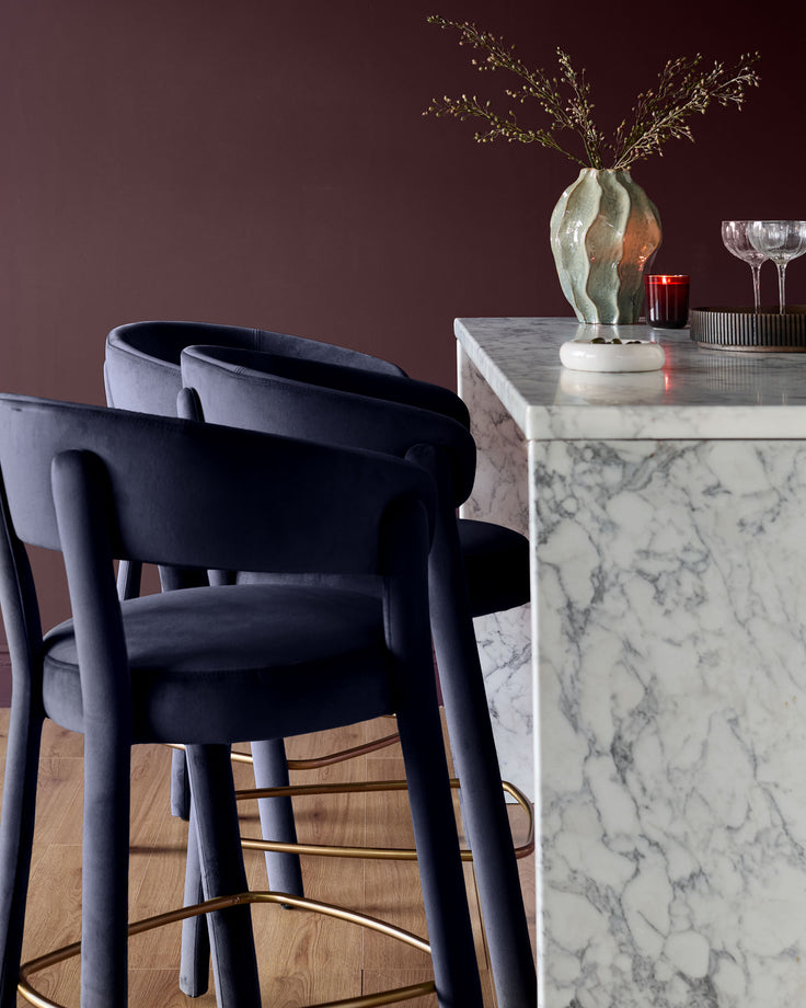 Anya bar stool in dark navy velvet with curved armrests and brass footrest, shown at marble counter.