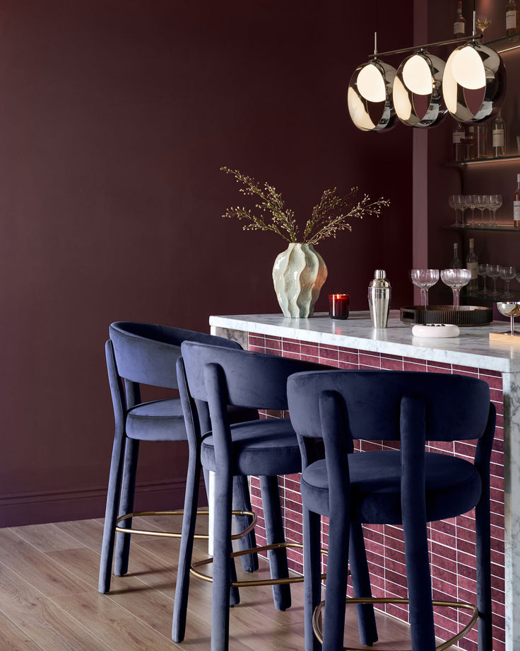 Anya bar stool in dark navy velvet with curved backrest and brass footrest, shown at marble bar counter.