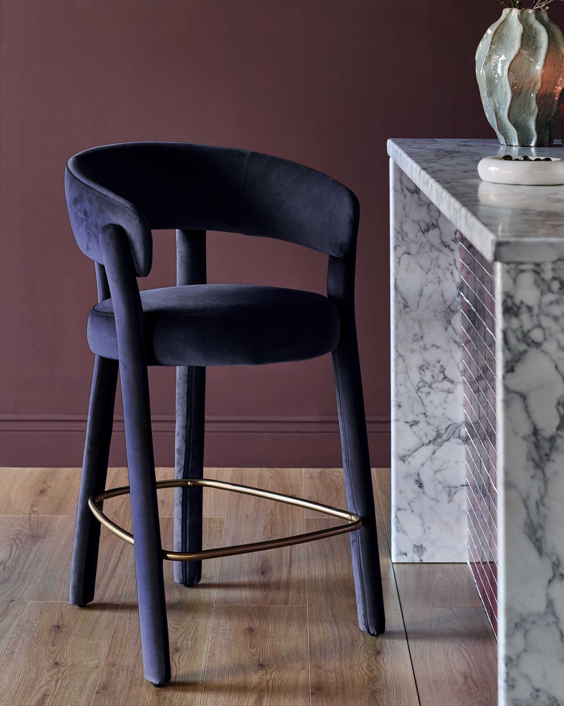 Anya bar stool in dark navy velvet with curved backrest and brass footrest, positioned next to marble table.