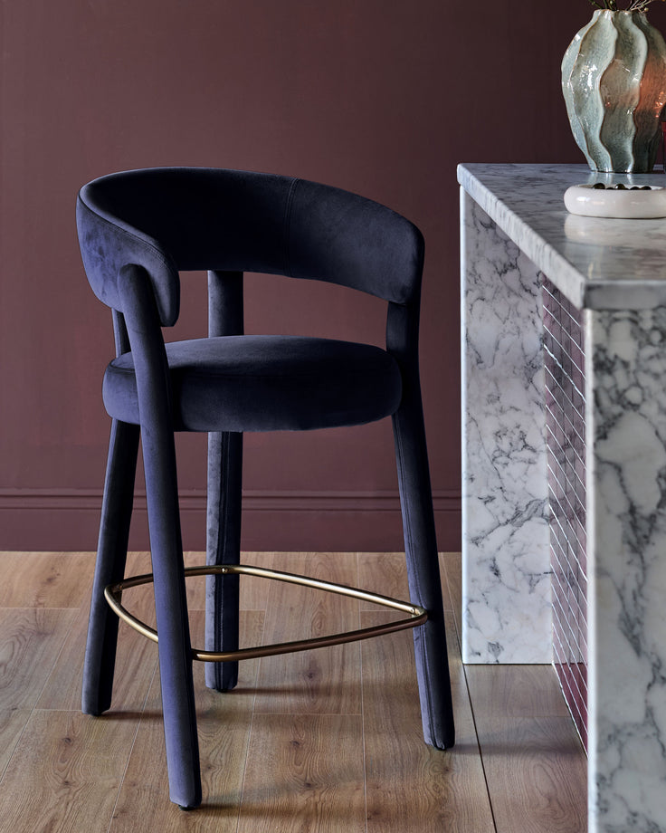 Anya bar stool in dark navy velvet with curved backrest and brass footrest, positioned next to marble table.