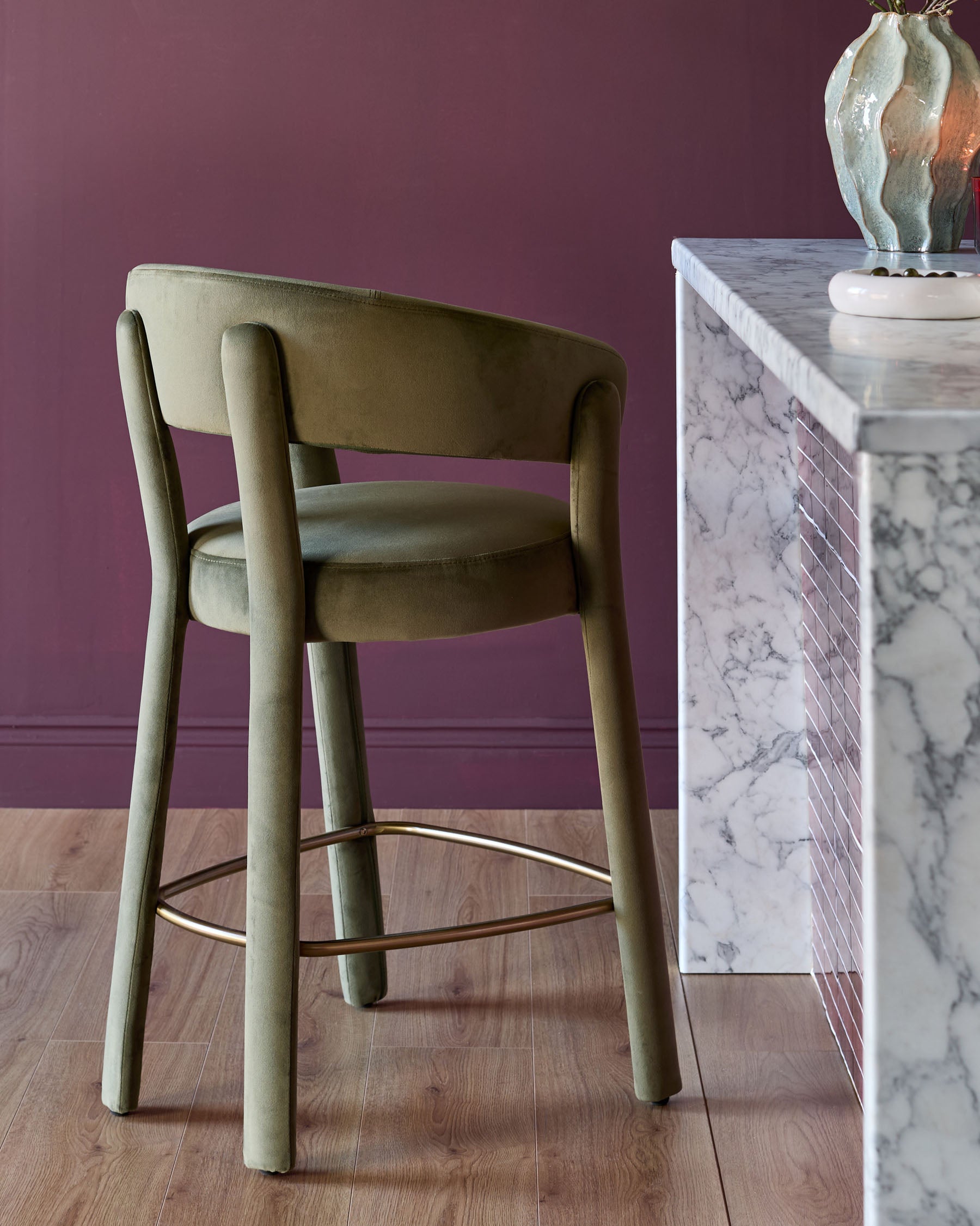 Anya bar stool in olive green velvet with curved backrest and wooden legs, paired with marble-topped console table.
