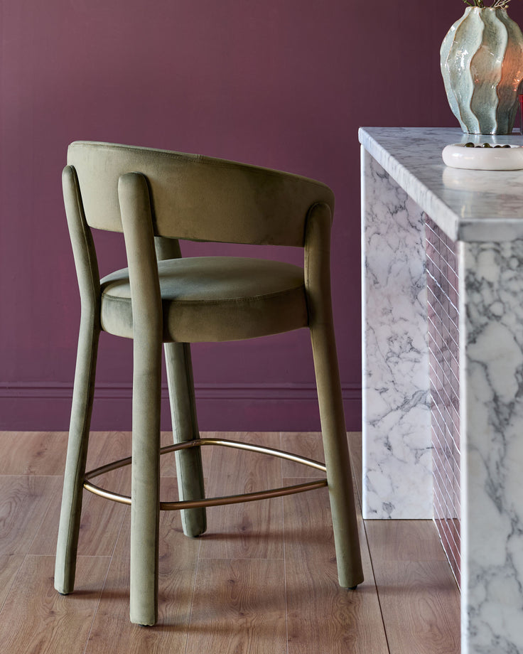 Anya bar stool in olive green velvet with curved backrest and wooden legs, paired with marble-topped console table.