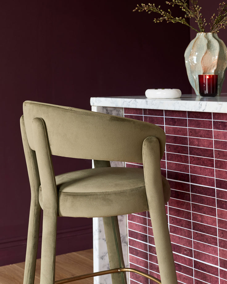 Olive green velvet bar stool with curved armrests and backrest, shown at an angle next to a marble and burgundy tile counter.