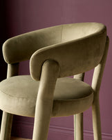 Olive green velvet bar stool with curved backrest and armrests, cylindrical legs, shown from side angle against purple background.