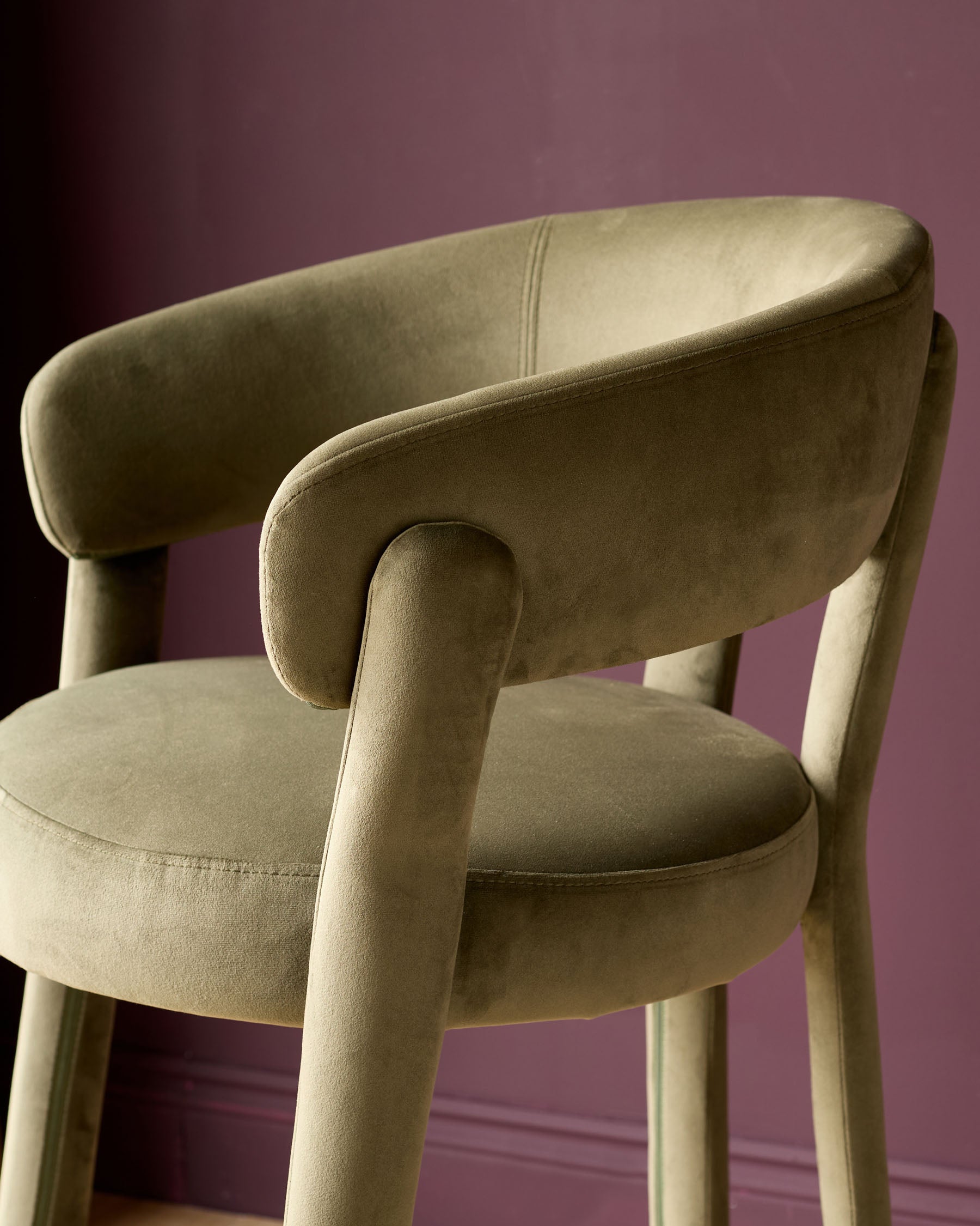 Olive green velvet bar stool with curved backrest and armrests, cylindrical legs, shown from side angle against purple background.