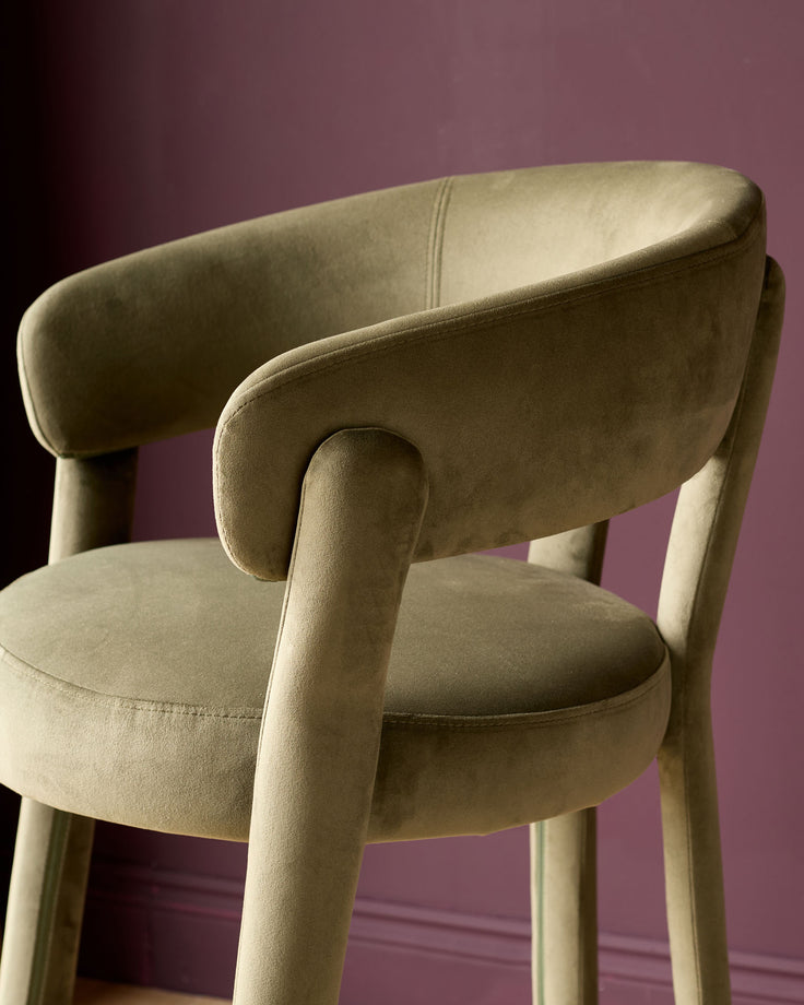Olive green velvet bar stool with curved backrest and armrests, cylindrical legs, shown from side angle against purple background.