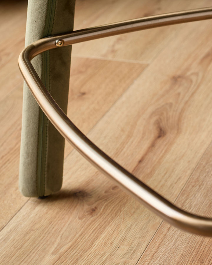 Anya bar stool in olive green velvet with brass curved footrest detail on wooden floor.