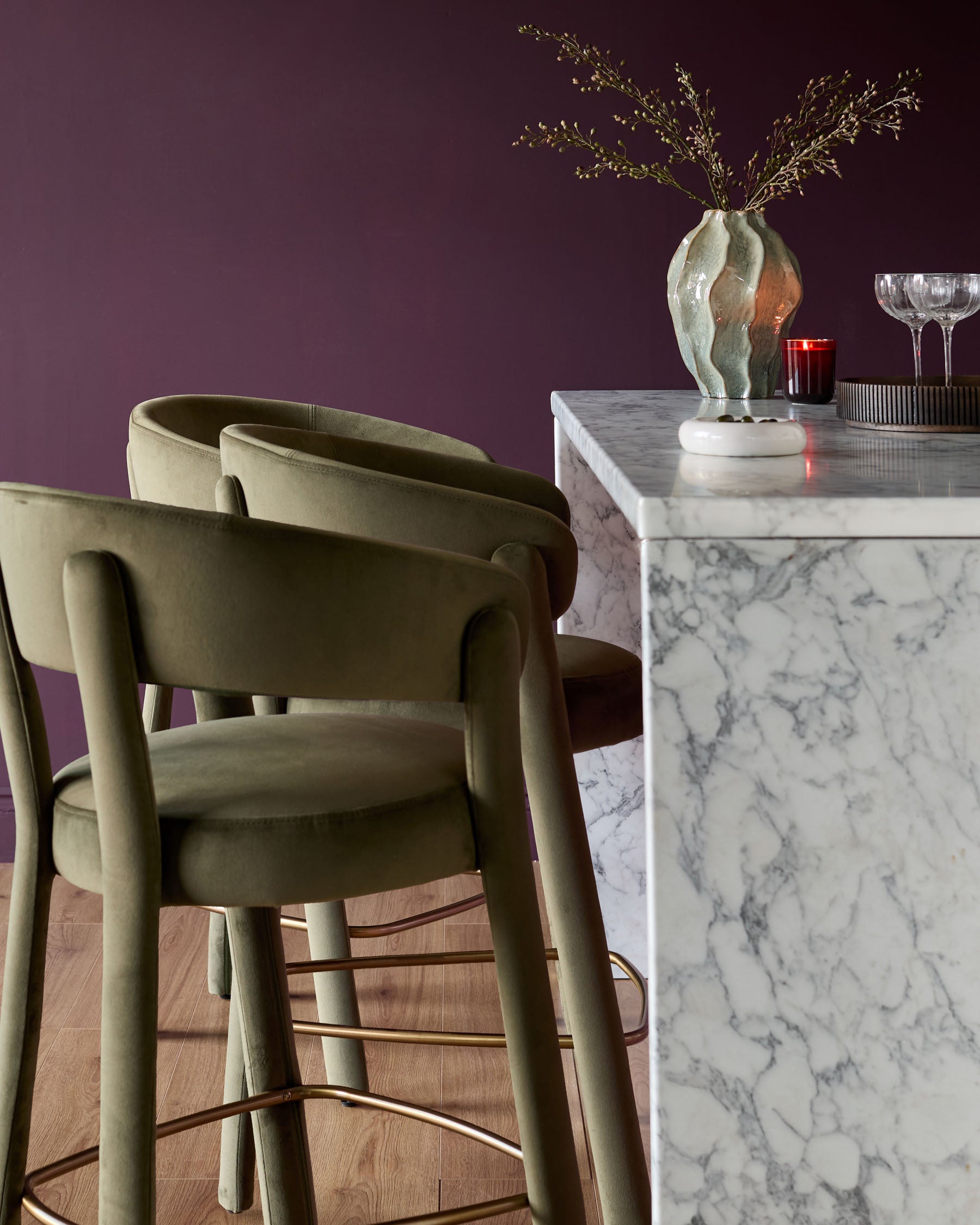 Olive green velvet bar stool with curved backrest and armrests, shown in a styled dining setting.