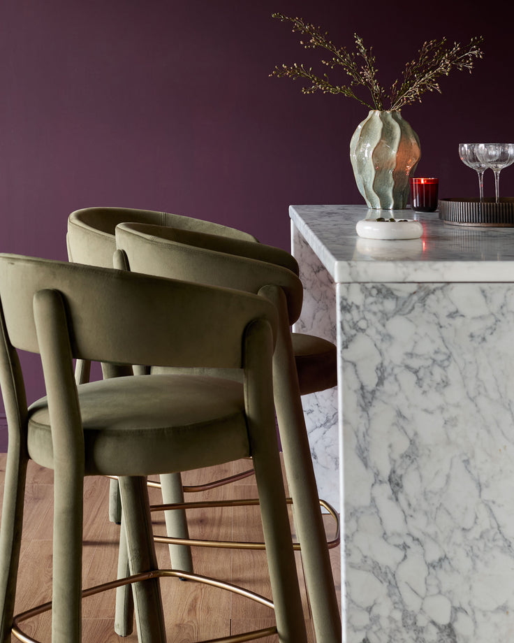 Olive green velvet bar stool with curved backrest and armrests, shown in a styled dining setting.