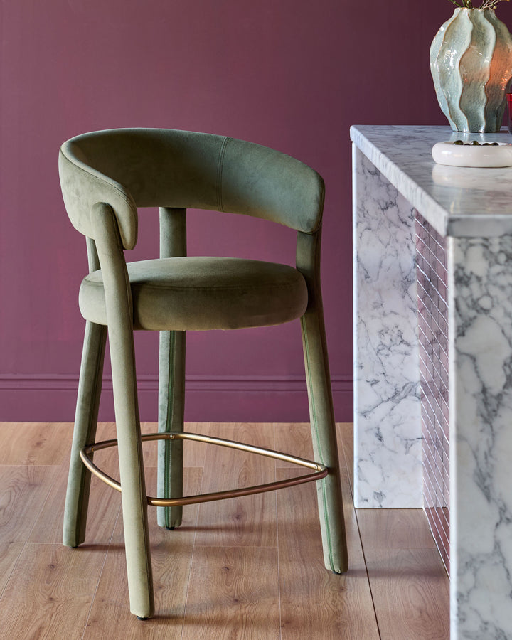 Anya bar stool in olive green velvet with curved backrest, wooden legs, and brass footrest detail.
