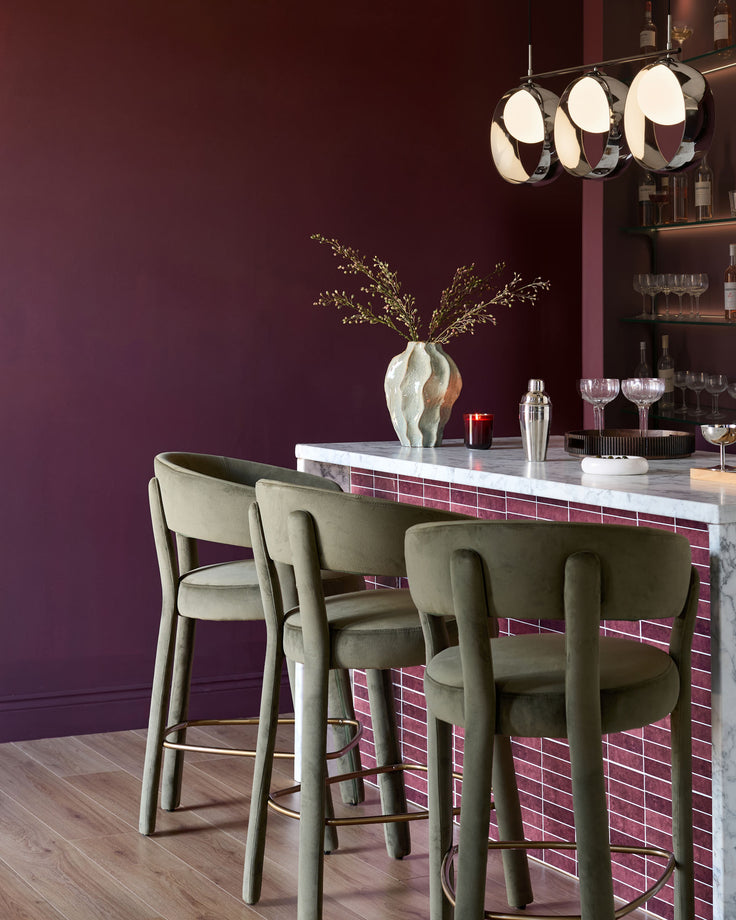 Olive green velvet bar stools with curved backrest and footrest, positioned at marble-topped bar counter.
