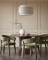 Dining area with round wooden table, striking base, four green velvet chairs and a tiered linen pendant light.