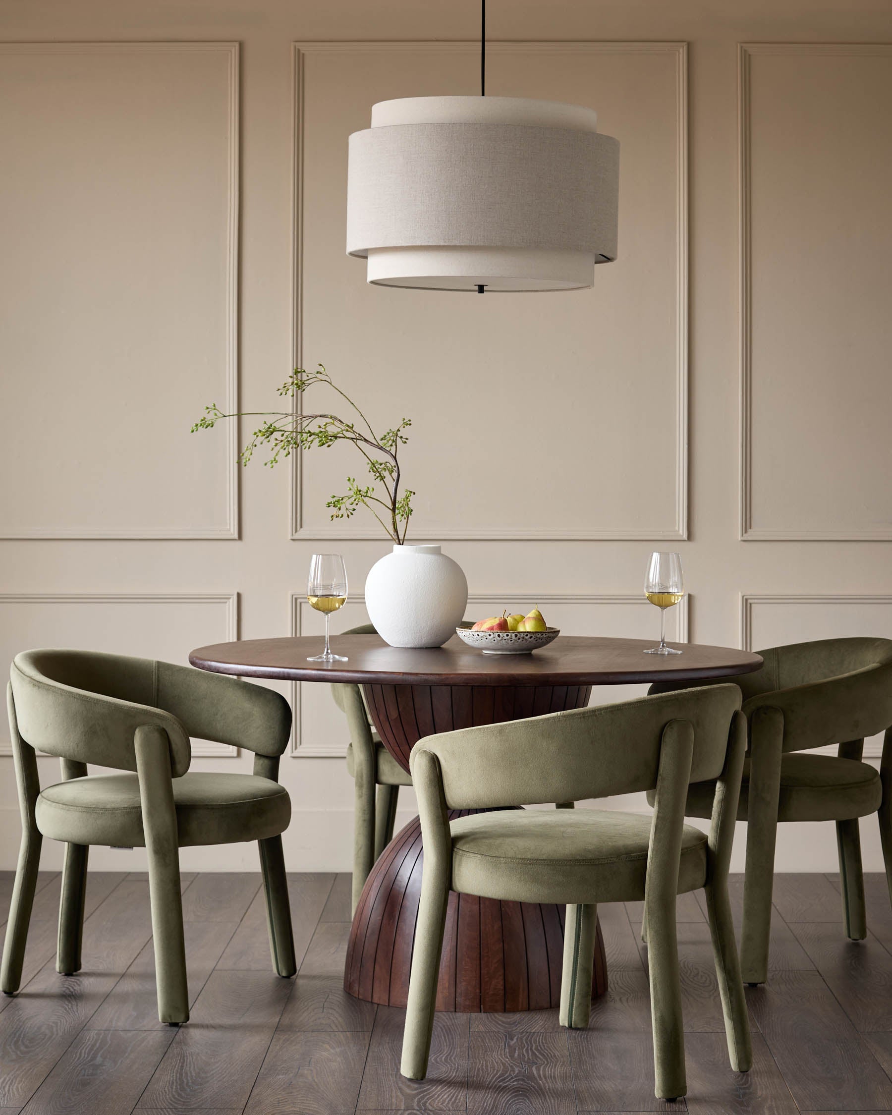Dining area with round wooden table, striking base, four green velvet chairs and a tiered linen pendant light.