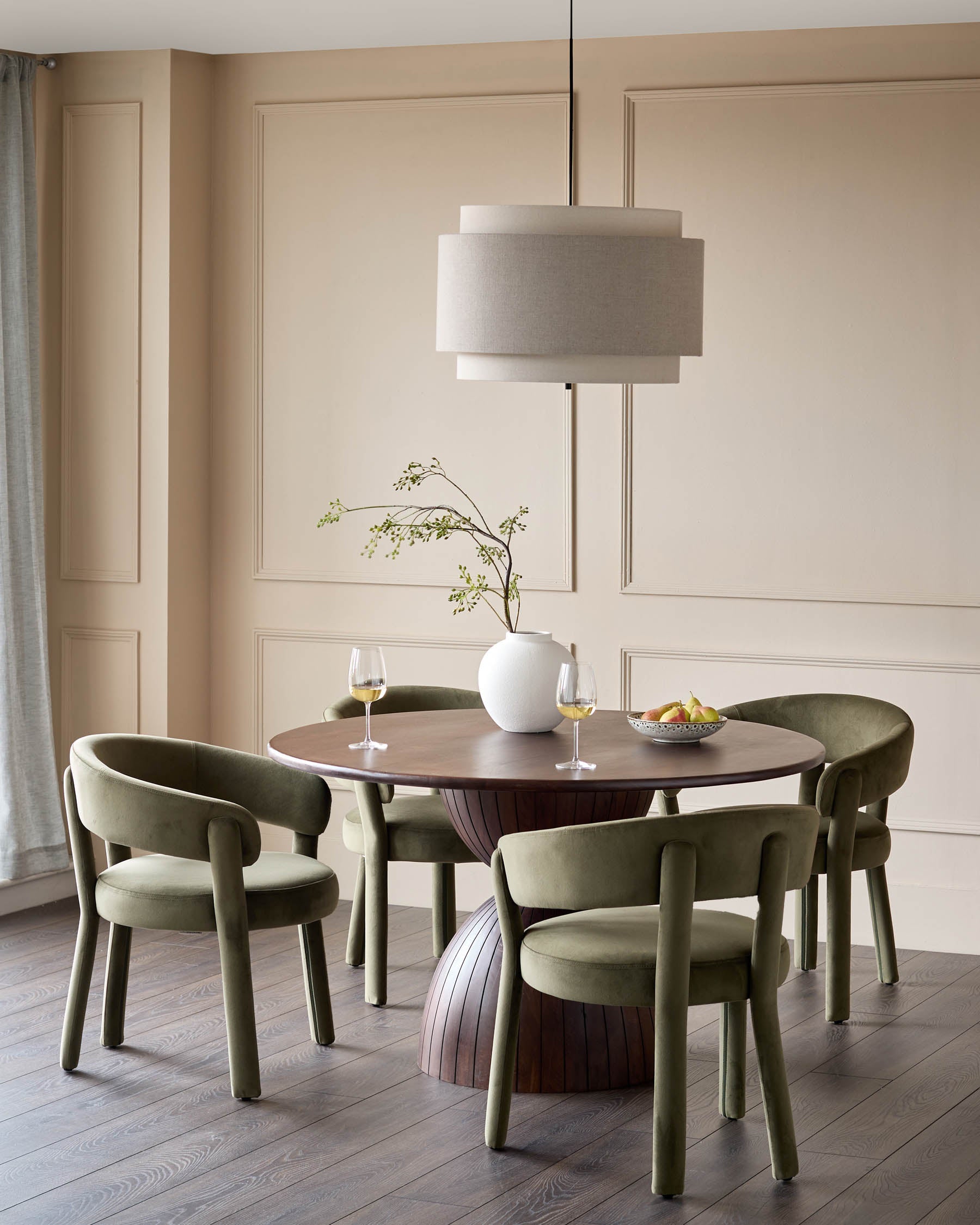A dining set with a round table, statement base, and green velvet dining chairs, under a tiered lamp.