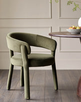 An olive green velvet chair sits beside a dark wood table with fruit, creating an inviting modern dining space.
