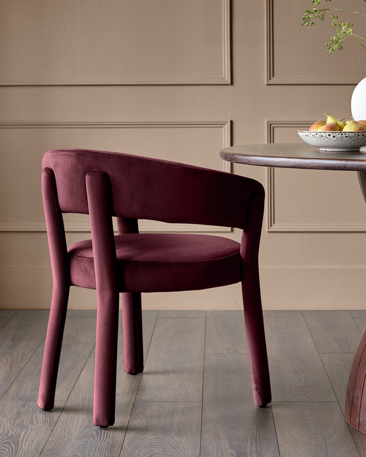 Plum velvet dining chair with curved backrest, cylindrical armrests, and tapered legs, shown in a styled room setting.