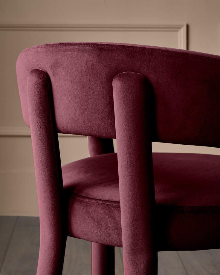 Anya dining chair in plum velvet, close-up of curved backrest and cylindrical armrests from side angle.