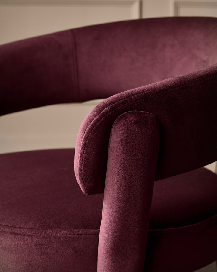 Anya dining chair in plum velvet, showing curved armrest and padded seat detail from side angle.