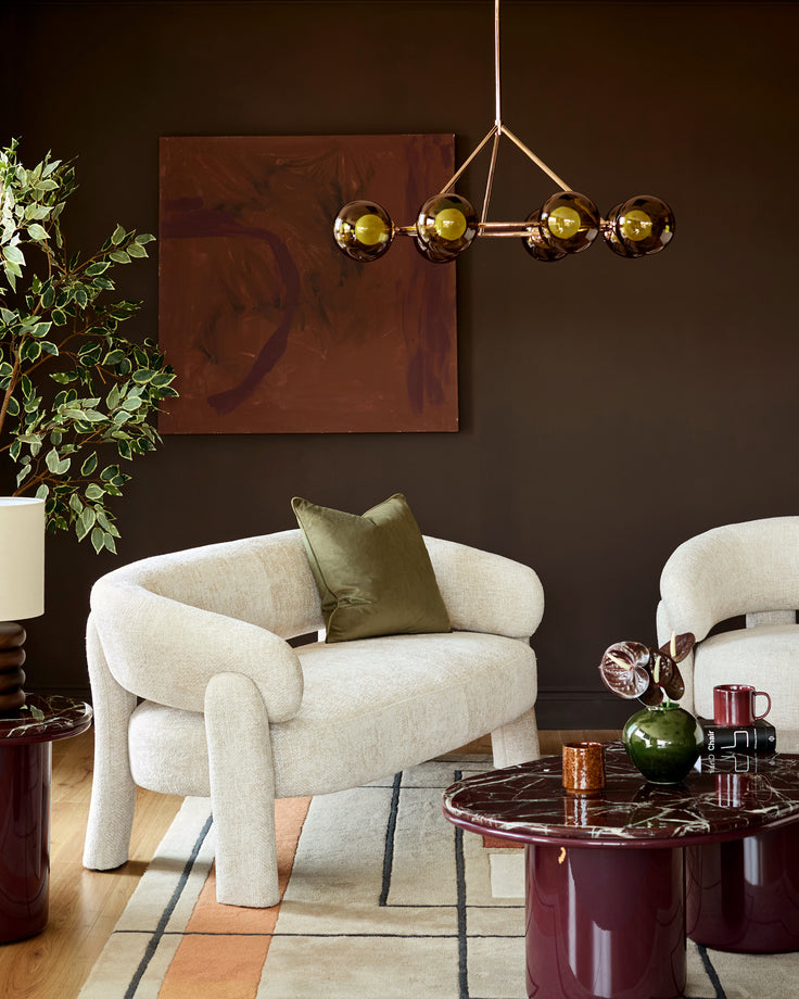 Anya accent sofa in natural fabric with curved arms and legs, styled in a modern living room with burgundy accents.