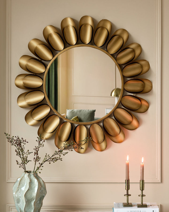 Gold round wall mirror with decorative petal-shaped frame, hung on beige wall with dried flowers and candles below.