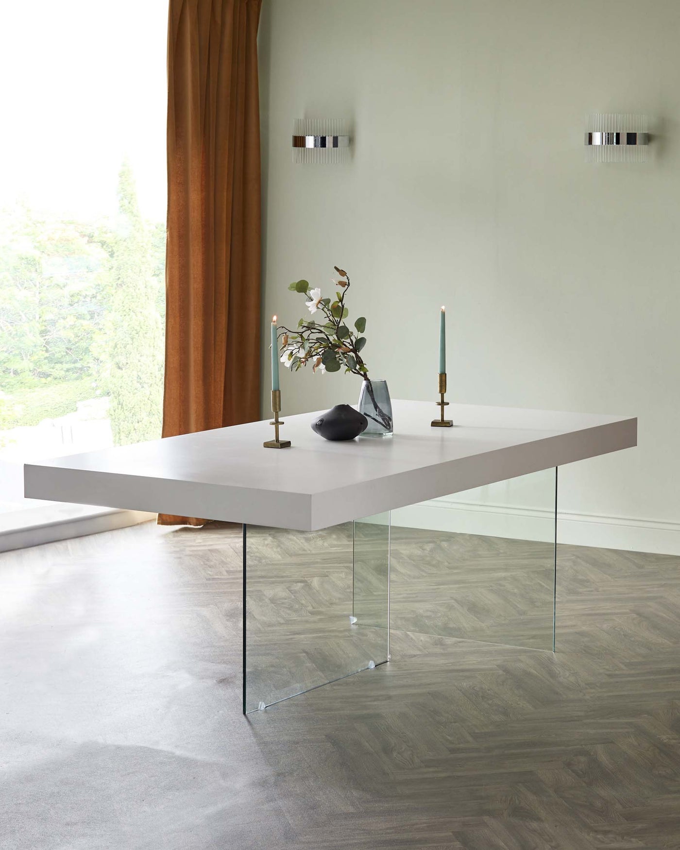 A modern minimalist dining table with a rectangular light grey top supported by two clear glass legs, set in a contemporary room with a view of greenery through a window.