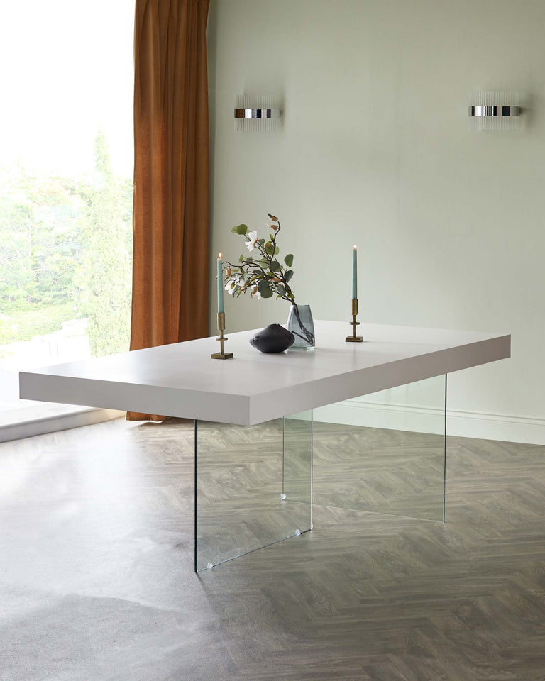 A modern minimalist dining table with a rectangular light grey top supported by two clear glass legs, set in a contemporary room with a view of greenery through a window.