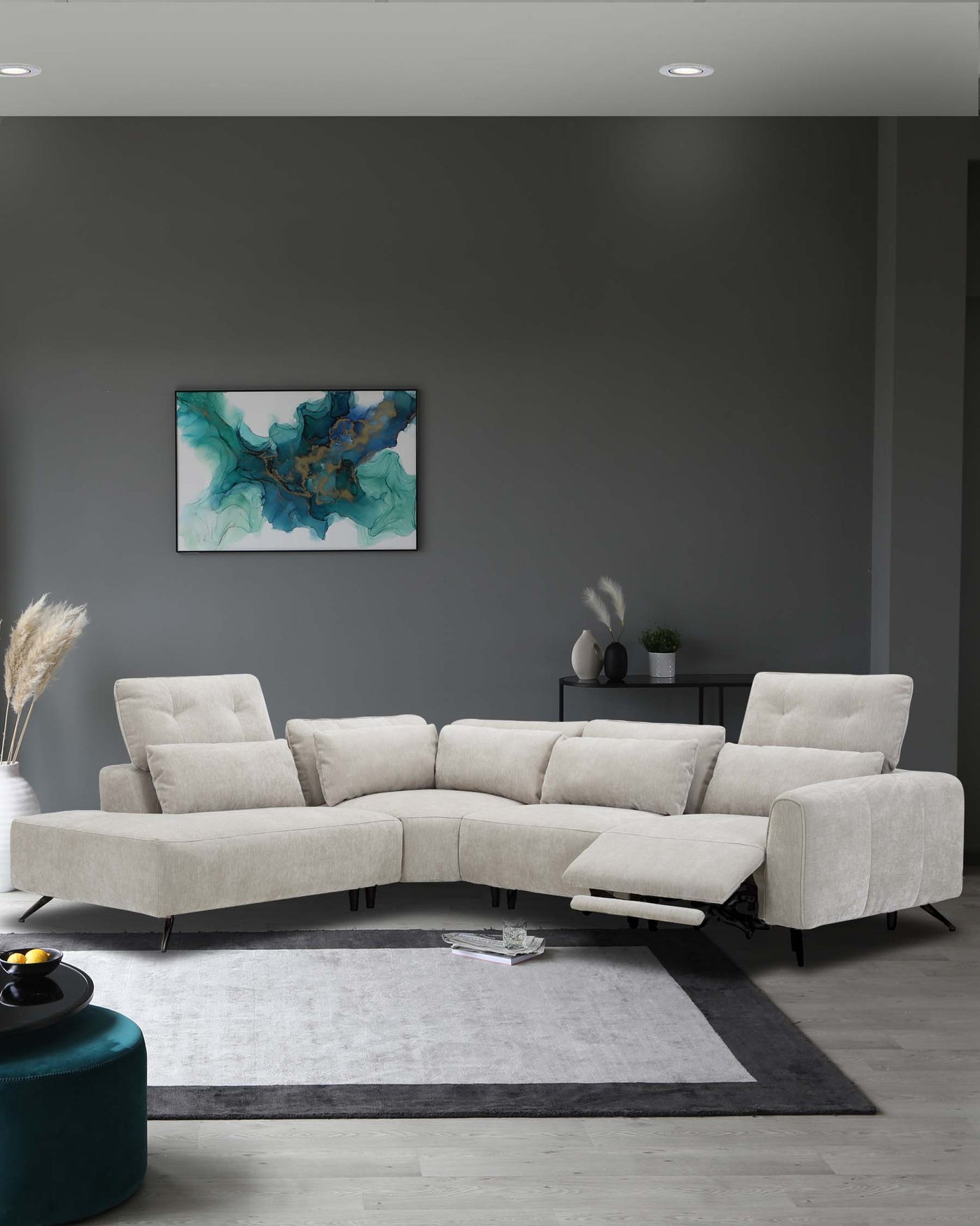 Modern L-shaped sectional sofa in light beige fabric with adjustable backrests and metal legs, positioned on a two-tone grey area rug. A round teal ottoman sits off to the side, with a sleek dark shelf unit in the background holding minimalistic decor.
