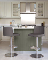 Two grey bar stools sit at a kitchen island with a white worktop and sage green panelling in a bright kitchen.