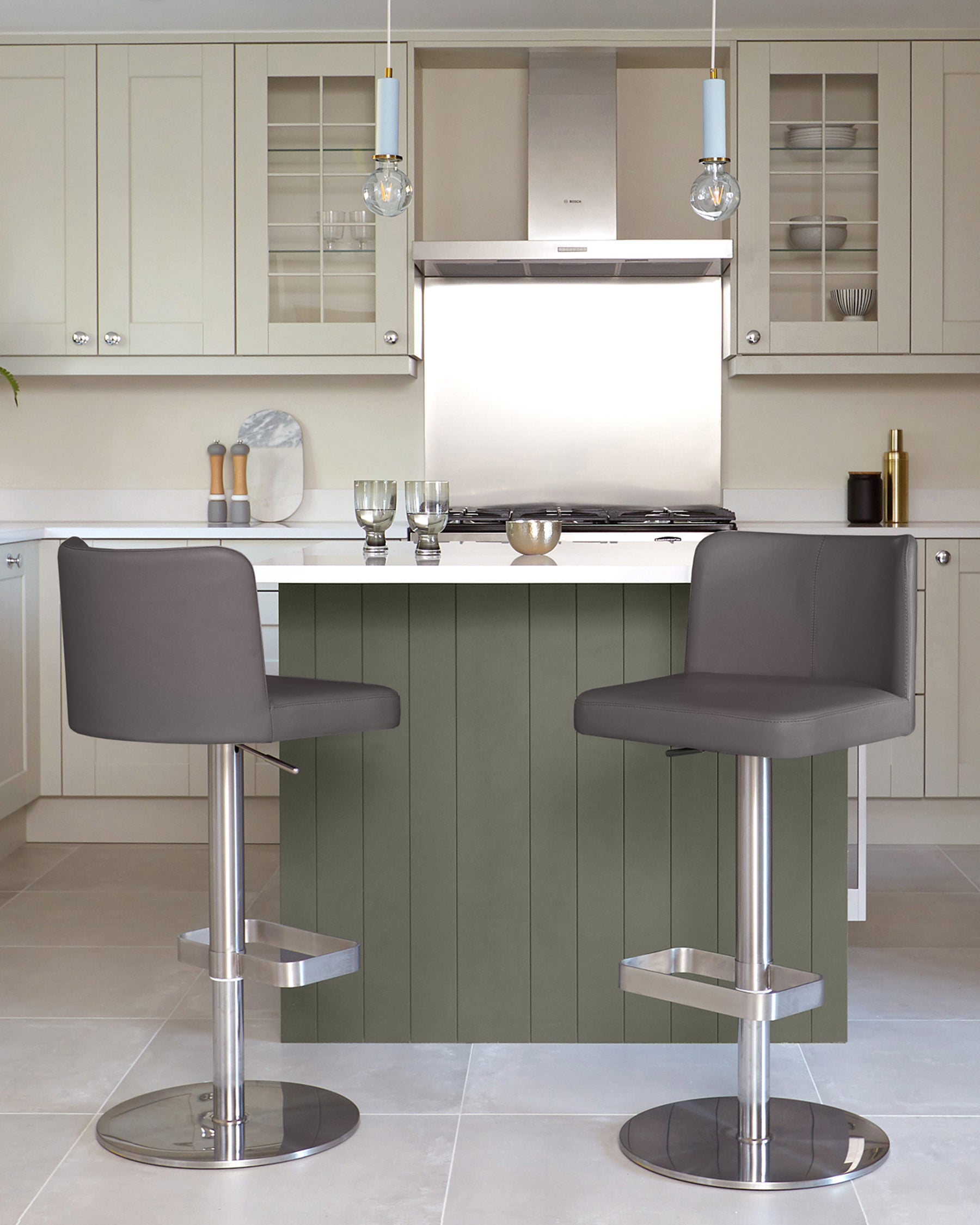 Two grey bar stools sit at a kitchen island with a white worktop and sage green panelling in a bright kitchen.