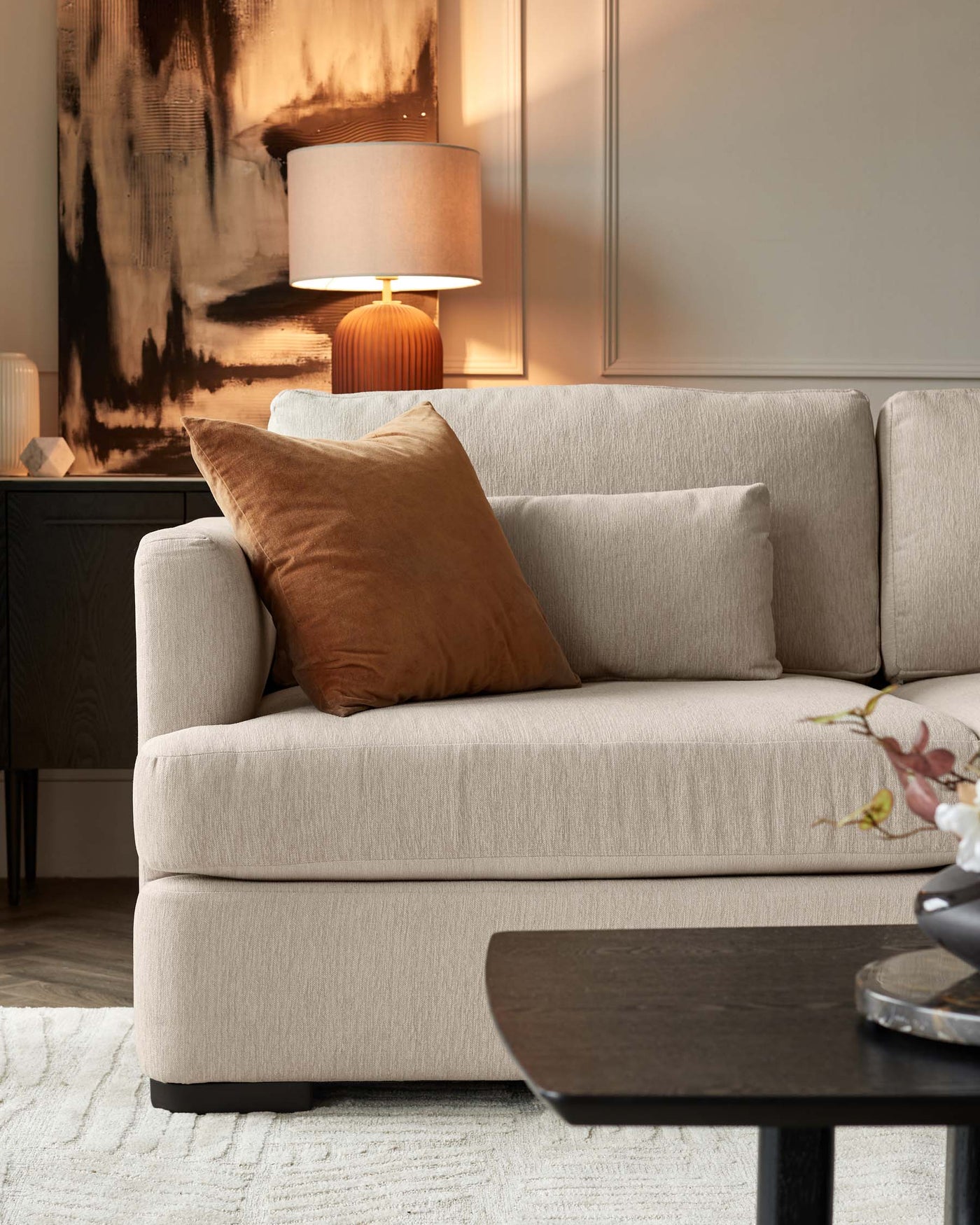A contemporary beige upholstered sofa with plush back cushions and a singular seat cushion, flanked by a dark wood side table with a ribbed texture base and a lamp with a warm-toned shade on top. In the foreground, a dark-stained, elliptical-shaped coffee table is partially visible, and a textured off-white area rug lies underneath. Decorative accents include neutral-toned pillows and an abstract painting in warm browns and blacks.