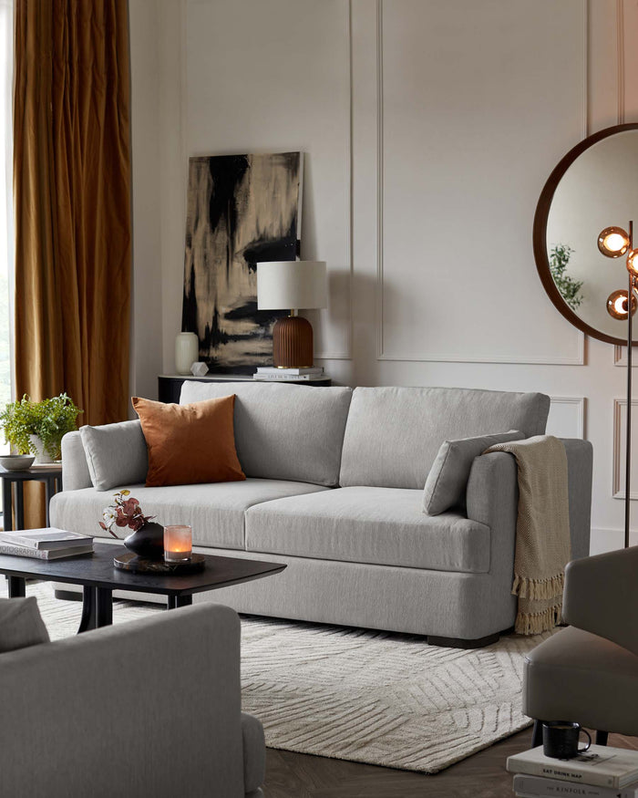 Elegant living room setup featuring a contemporary light grey upholstered sofa with plush cushions and a matching armchair. A sleek, black round coffee table sits on a textured off-white area rug, complemented by a small side table with a modern lamp atop.