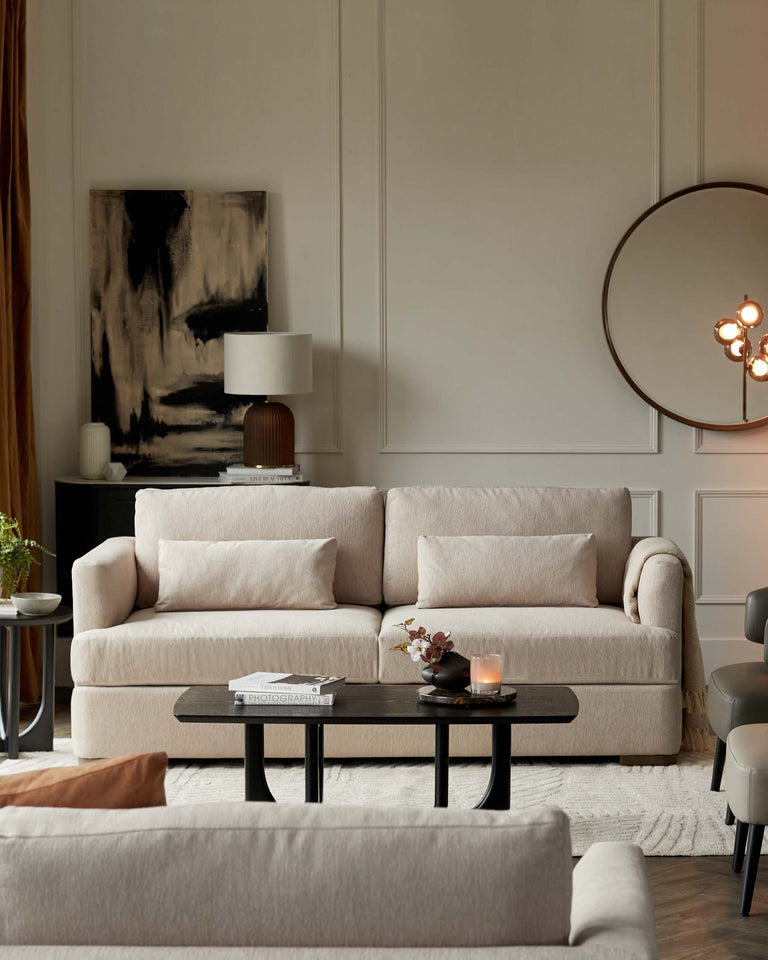 Elegant living room interior featuring a plush, light taupe three-seat sofa with soft fabric upholstery and matching side armrests. In front of the sofa is an oval-shaped, black coffee table with a sleek design. A smaller side table with a dark surface and thin legs is to the side, supporting a lamp with a unique ribbed base and a white shade. Completing the scene is a beige accent armchair with a modern silhouette to the right, harmonizing with the room's warm neutral tones and sophisticated ambiance.
