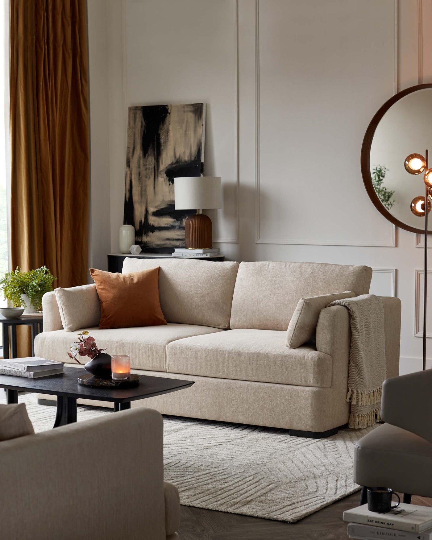 Contemporary beige three-seater sofa with clean lines and plush cushions, accented with a burnt orange throw pillow. In front, a low-profile oval-shaped coffee table in dark wood. To the side, a small round end table with a potted plant complements the space. A textured off-white area rug anchors the seating arrangement, while an armchair facing the sofa completes the setting. Elegant drapery, modern art, and a round wall mirror enhance the room's sophisticated aesthetic.