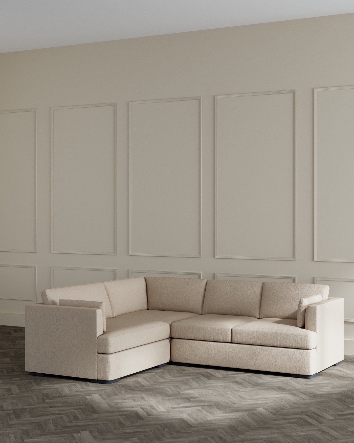Modern beige L-shaped sectional sofa with a minimalist design and clean lines, set against a neutral wall with decorative panelling, positioned on a dark herringbone-patterned floor.