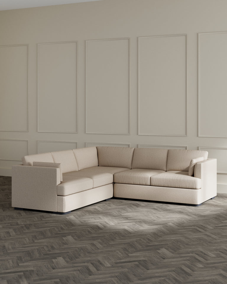 Modern L-shaped sectional sofa in a light beige fabric with clean lines and comfortable cushions displayed in a minimalist room with herringbone wood flooring and elegant panelling on the walls.