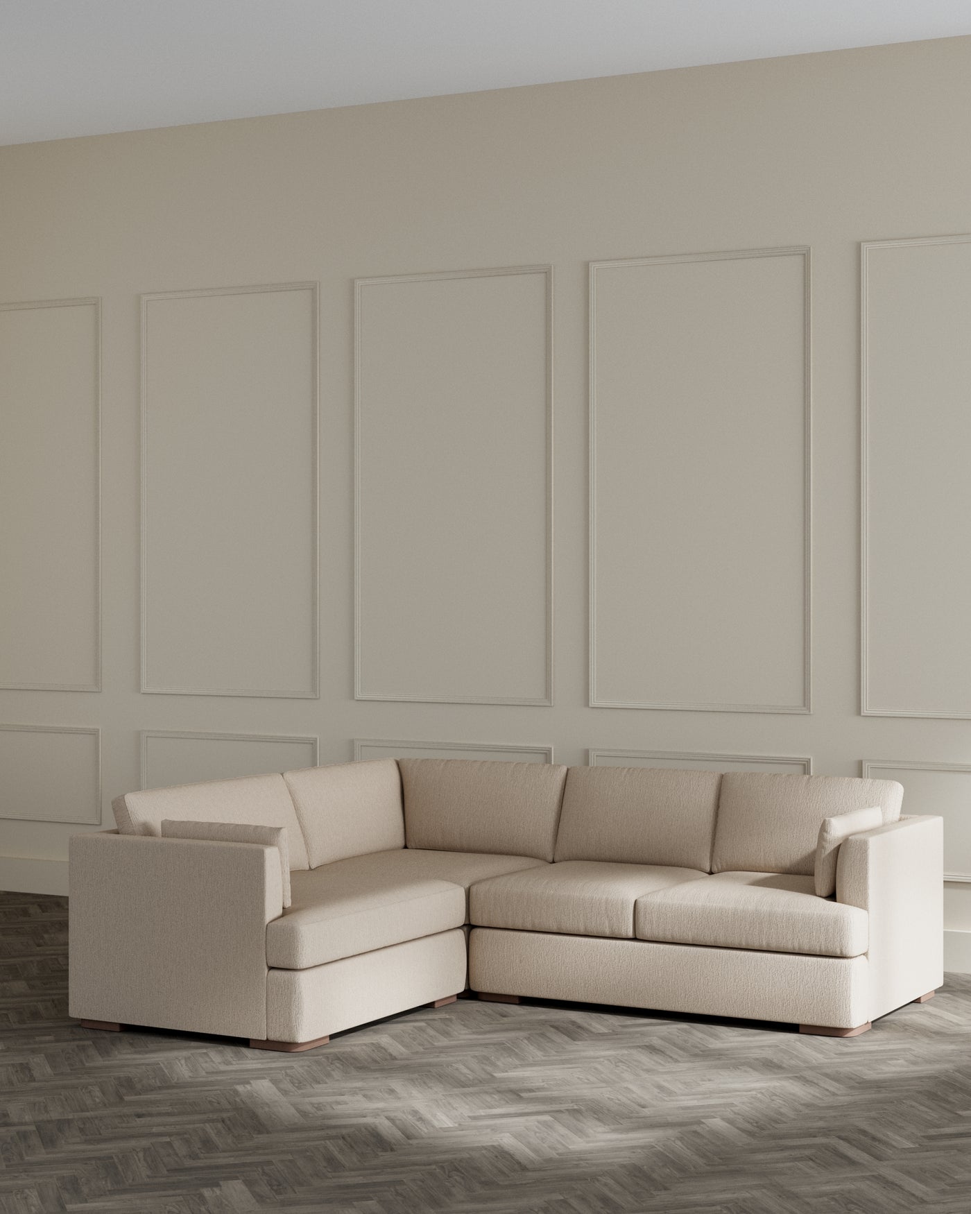 Modern beige L-shaped sectional sofa with clean lines and plush cushions, positioned on a herringbone-pattern hardwood floor against a panelled wall.