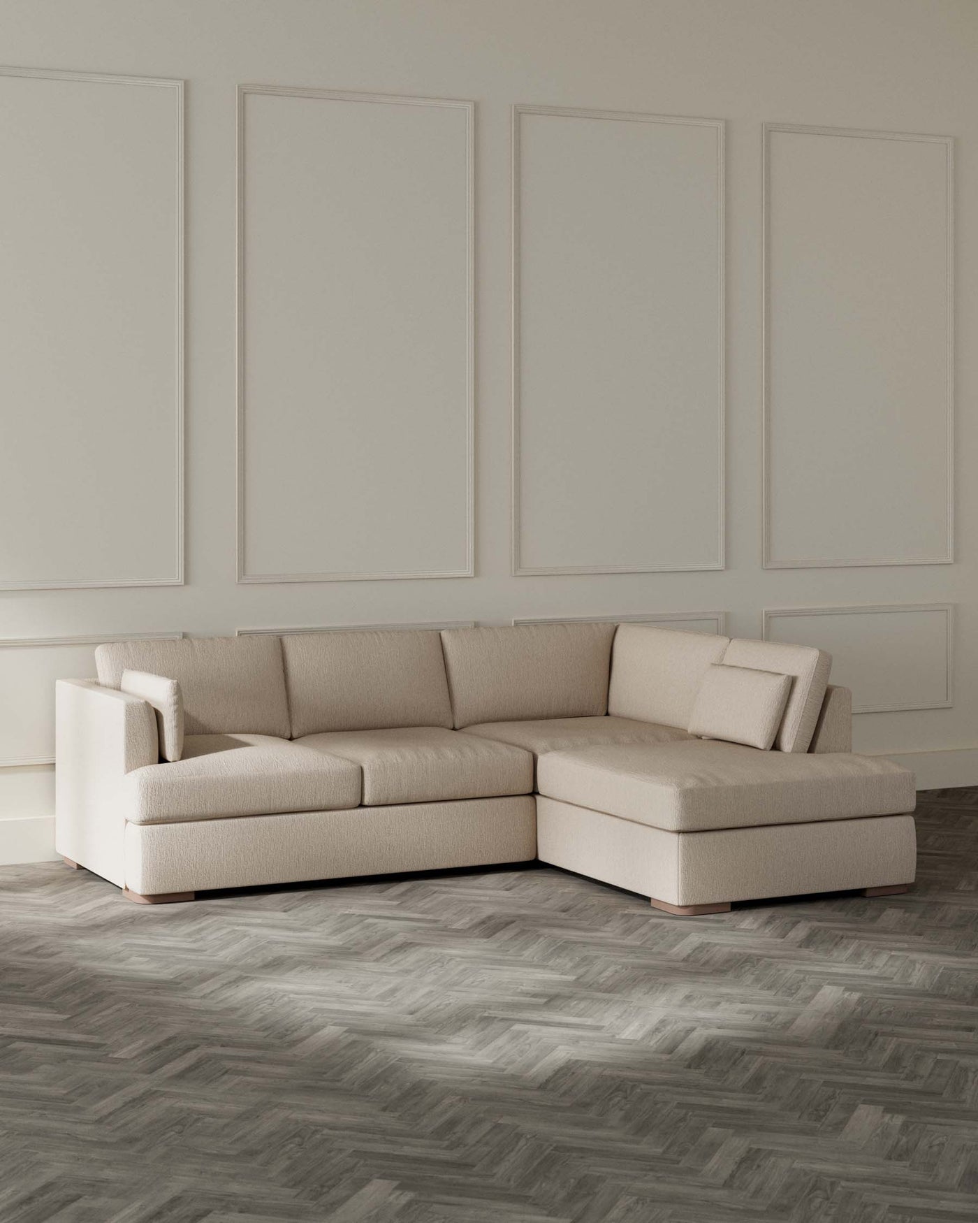 Contemporary L-shaped sectional sofa with a light beige fabric upholstery and clean lines, featuring plush back cushions and a chaise lounge extension, set on a dark herringbone-patterned floor with a neutral wall punctuated by decorative mouldings in the background.