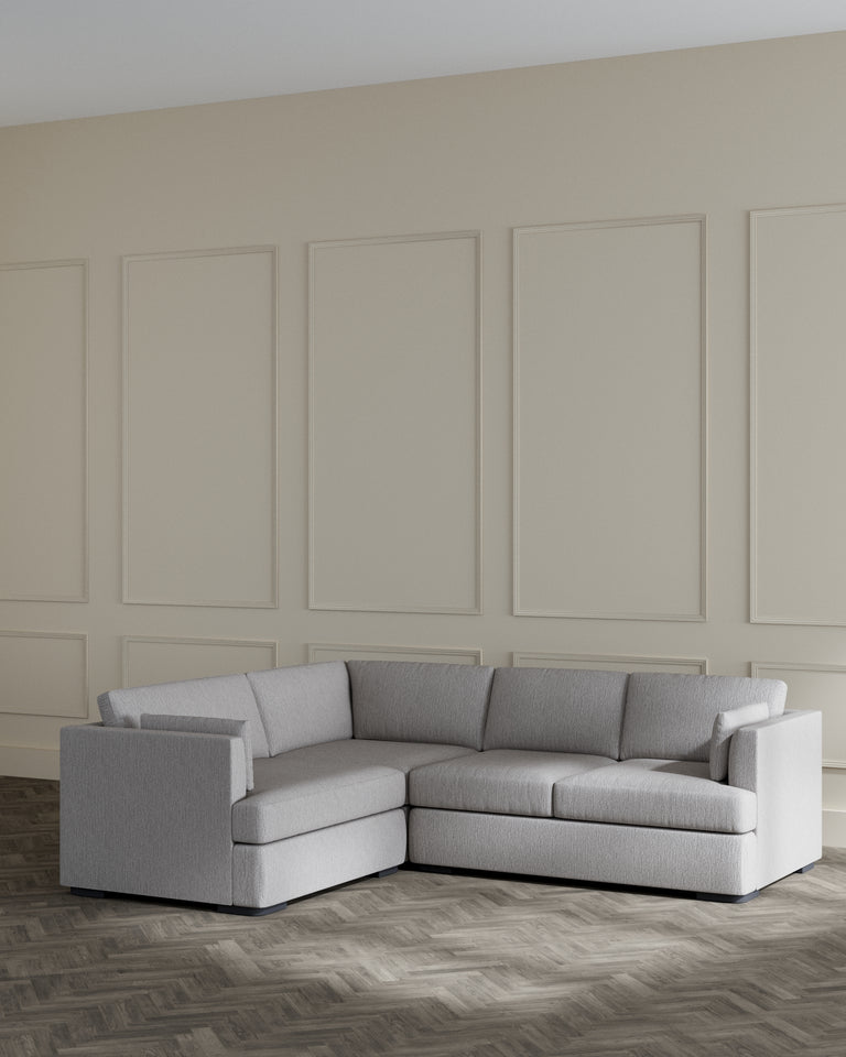 Modern L-shaped sectional sofa with a light grey fabric upholstery and clean, simple lines, featuring plush back cushions and a chaise lounge on the left side. The sofa is set against a neutral wall with elegant panelling and rests on a herringbone patterned wooden floor.