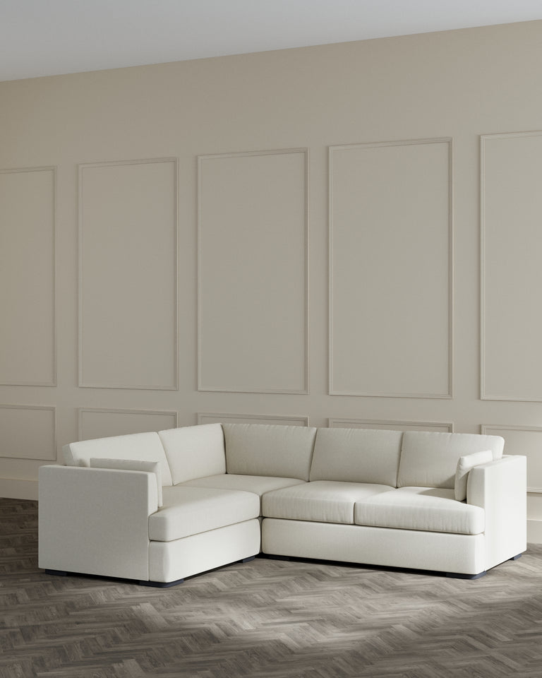 A modern L-shaped sectional sofa in a light cream fabric, featuring clean lines and a simple, elegant design with plush cushions and a low-profile silhouette.