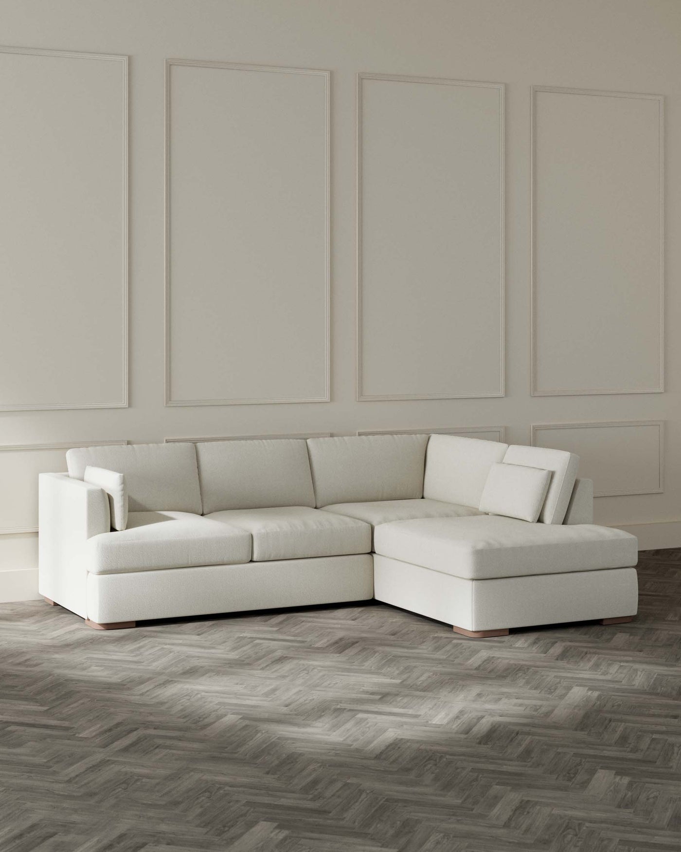 A modern L-shaped sectional sofa in a light cream fabric with a chaise on the left and a minimalist design, set against a neutral wall with decorative panels and on a herringbone-patterned wooden floor.