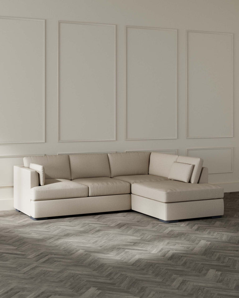 Beige L-shaped sectional sofa with chaise lounge on a herringbone-patterned wooden floor against a wall with decorative panelling.
