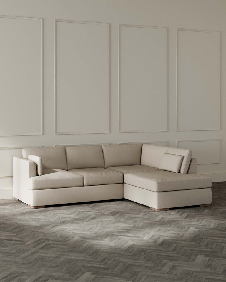 Elegant modern beige sectional sofa with chaise lounge on a herringbone wood floor against a wall panelled with rectangular white moulding.