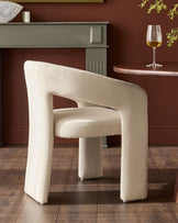 Cream fabric dining chair with curved, rounded edges and padded arms, shown from a three-quarter angle in a styled room setting.
