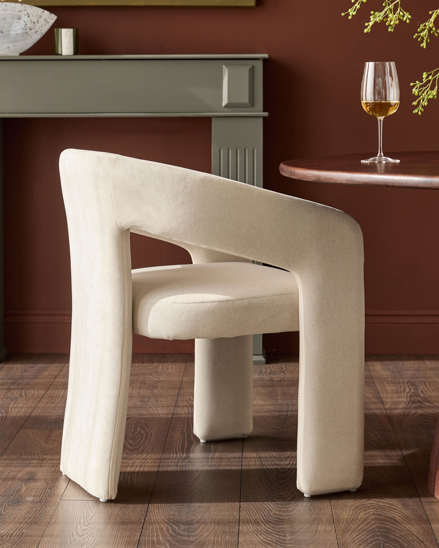 Cream fabric dining chair with curved, rounded edges and padded arms, shown from a three-quarter angle in a styled room setting.