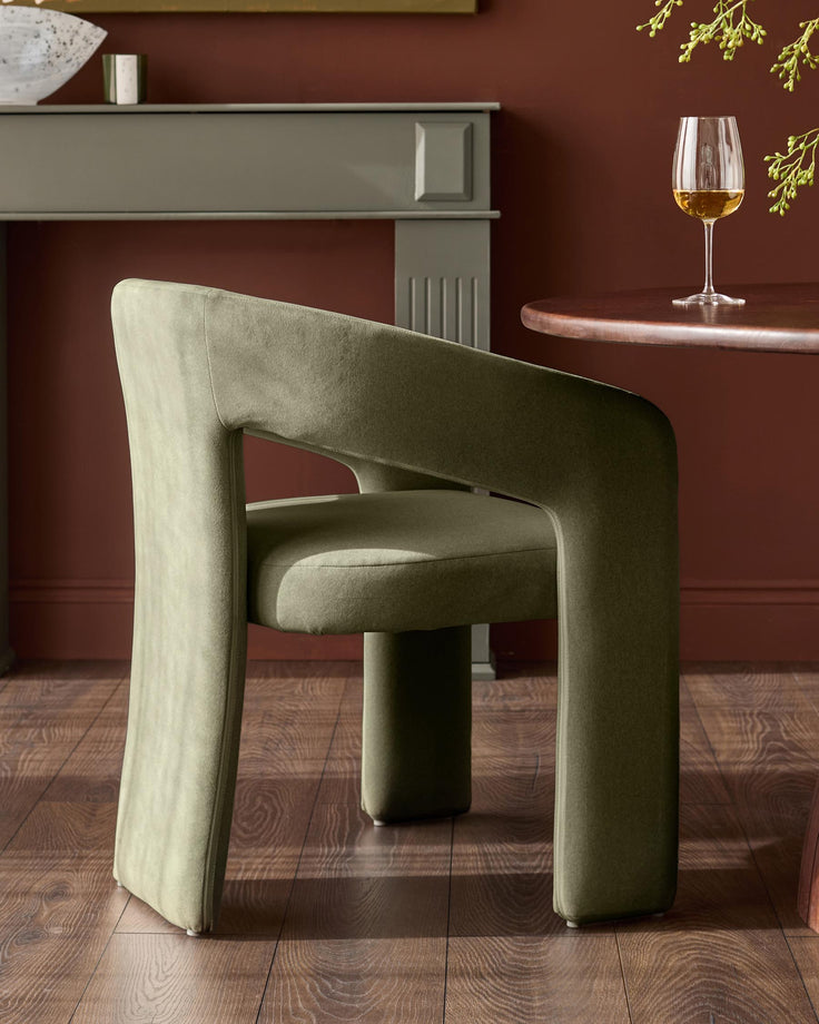 Asher dining chair in olive green fabric, angled side view showing curved armrests and cylindrical base in modern interior setting.