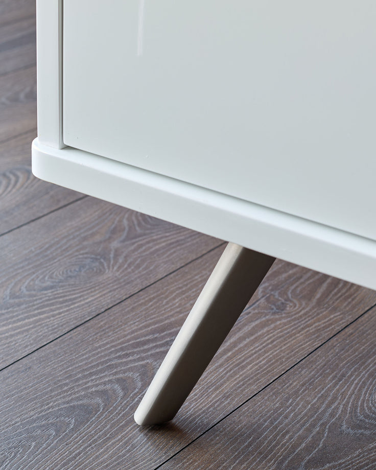 White compact sideboard with glossy finish, shown at an angle on dark wood flooring, featuring a white cylindrical leg detail.