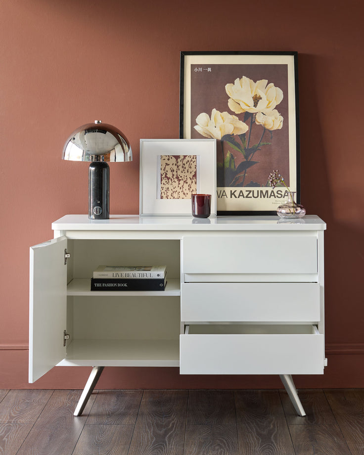 White gloss compact sideboard with open door revealing shelving, three drawers, and tapered legs against a terracotta wall.