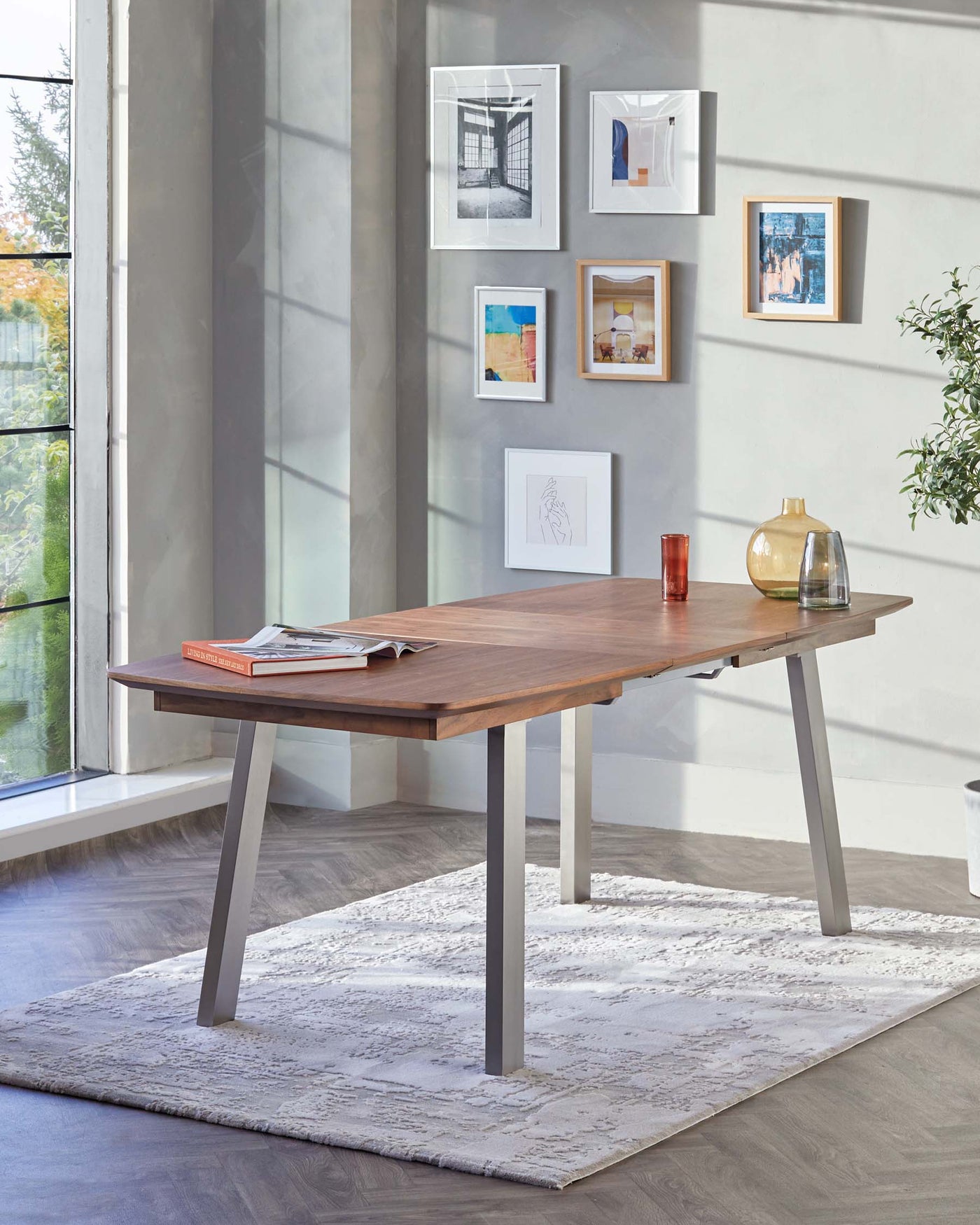 Modern wooden dining table with a warm, natural wood finish and contrasting sleek, grey metal legs, presented in a bright room with natural light. A minimalistic design aesthetic is emphasized by a few decorative items on the tabletop and framed artworks on the wall. The table is situated on a textured white area rug that softens the room's ambiance.