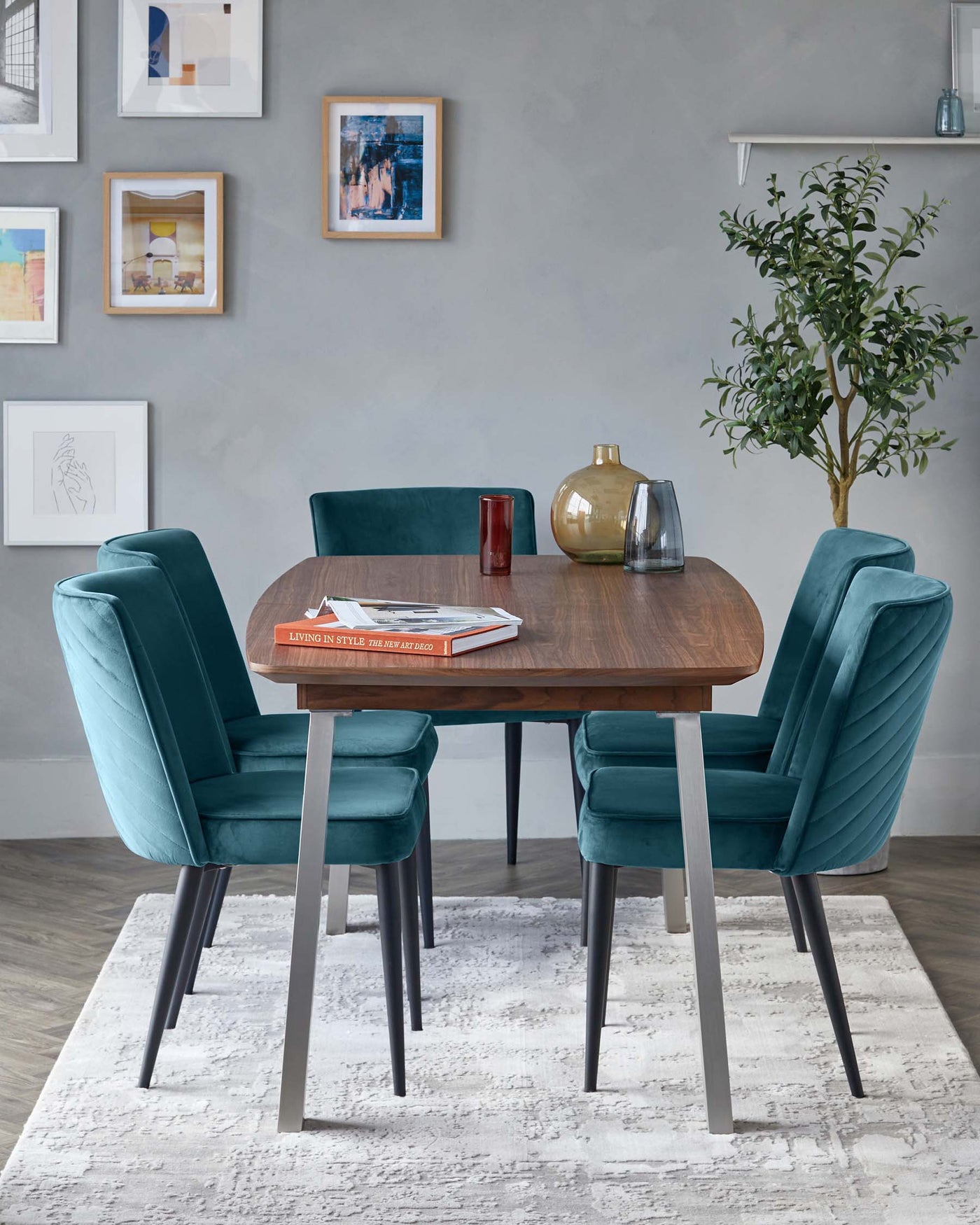 Elegant dining room setup featuring a contemporary round, walnut finish table with tapered metal legs, surrounded by four plush teal velvet chairs with channel tufting and metallic accents on the legs, arranged on a white and grey patterned area rug.
